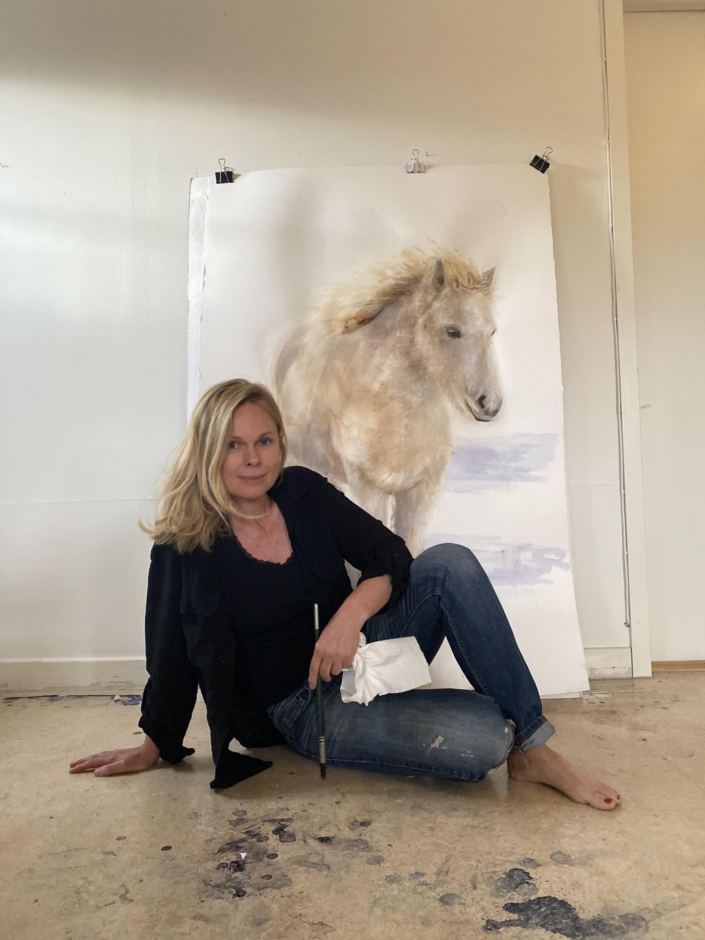 Anette Gustafsson, akvarellisti istuu lattialla valoisassa huoneessa, taustalla akvarelliteos missä näkyy valkoinen hevonen. Konstrundan, Taiteen perjantai tehtävä Fantastinen Akvarelli